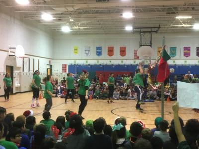 photo of teachers playing volleyball against students in the gym with student body seated on floor watching and cheering