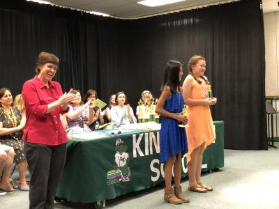 photo of students and teacher on stage next to table of trophies